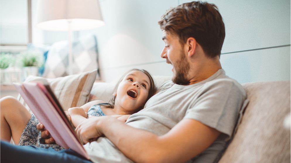 Ein Vater sitzt mit seiner Tochter auf der Couch und liest. Beide sind fröhlich. Kinder, die gut lesen können, profitieren von vielen Vorteilen: in der Schule, im Beruf und im ganzen Leben. So könnt ihr eure Kinder beim Lesenlernen fördern. | Bild: Wochit/ Getty Images