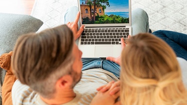 Paar sitzt auf einem Sofa und plant seinen Urlaub | Bild: mauritius images / György Barna / Alamy / Alamy Stock Photos
