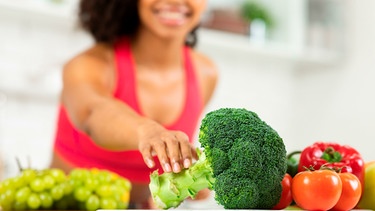 Eine Frau greift in ihrer Küche zu einem Brokkoli, daneben liegen Tomaten, Paprika und Weintrauben | Bild: mauritius images / Prostock-studio / Alamy / Alamy Stock Fotos