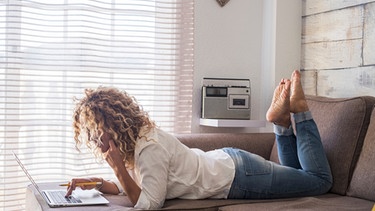 Eine Frau liegt entspannt auf dem Sofa, blickt in den Laptop und hört dabei Radio. | Bild: mauritius-images